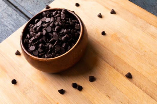 High angle view of chocolate chips in wooden bowl on cutting board. unaltered, sweet food and unhealthy eating concept.