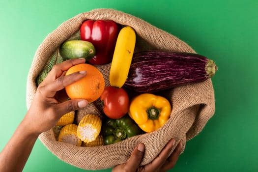 Directly above view of mixed race man hand holding fresh orange fruit over vegetables in burlap sack. unaltered, food, healthy eating, organic concept.