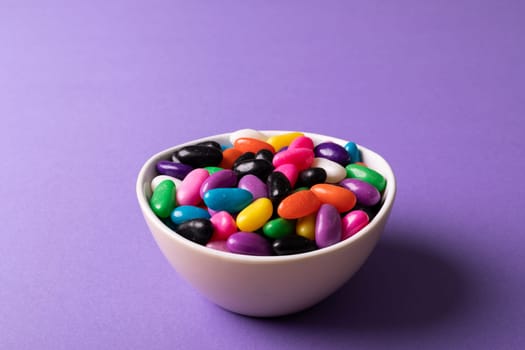 Close-up of multi colored candies in white bowl on purple background with copy space. unaltered, sweet food and unhealthy eating concept.