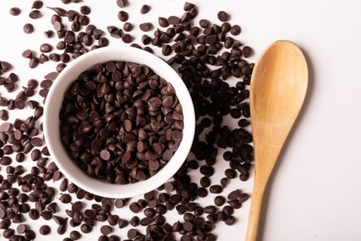 Overhead view of fresh chocolate chips with bowl and wooden spoon on white background. unaltered, sweet food and unhealthy eating concept.