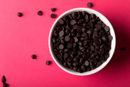 Overhead view of fresh chocolate chips in bowl over pink background. unaltered, sweet food and unhealthy eating concept.