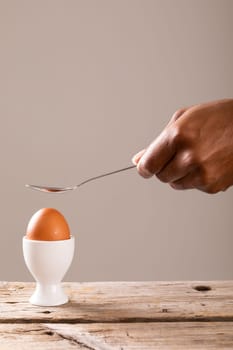 Cropped hand holding spoon over boiled brown egg in cup against gray background with copy space. unaltered, food, healthy eating and organic concept.