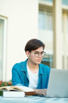Young male college student with eyeglasses and in casual clothings sitting outdoor to study, read books, and type using laptop for school project and research. Education and E-learning concept.