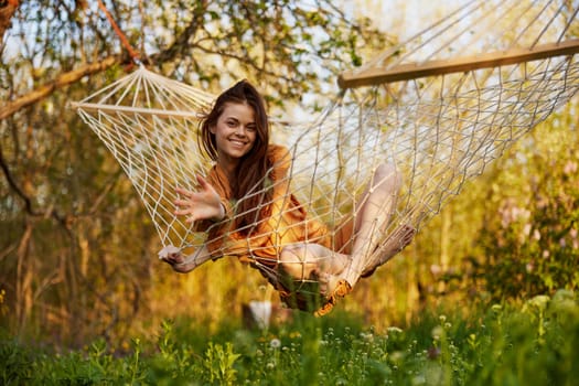 a joyful woman with long hair is lying in a hammock in an orange dress and happily smiling at the camera. A photo taken on the street on the theme of recreation in the country. High quality photo