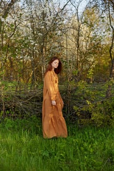 an elegant, sophisticated woman poses relaxed standing near a wicker fence at the dacha in a long orange dress looking to the side. Vertical photography in nature. High quality photo