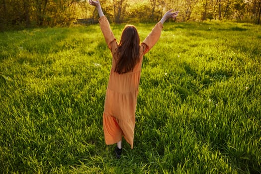 an attractive, slender, red-haired woman stands in a wide, green field during sunset in a long orange dress enjoying unity with nature and relaxation raising her arms to the sides while standing with her back to the camera. High quality photo