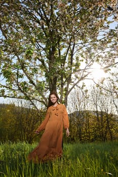 a sweet, attractive woman with long red hair stands in the countryside near a flowering tree in a long orange dress and looks at the camera with a slight smile on her face. High quality photo