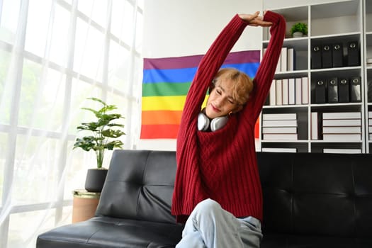 Carefree young gay man relaxing on couch at home and stretching arms. LGBTQ, people lifestyle, leisure concept.