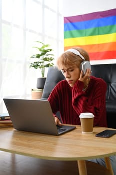 Focused young Asian gay man student in headphone looking at laptop screen watching tutorial, lecture or studying online at home.