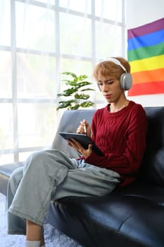 Portrait of casual teenage gay man wearing headphone using digital tablet on couch in living room with pride rainbow flag on wall.