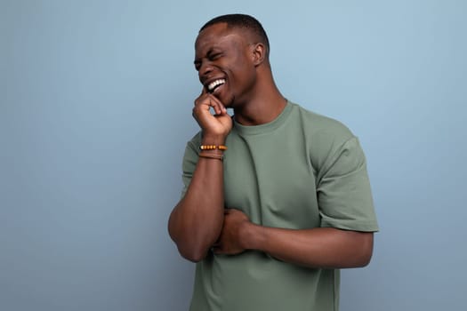 shy young american man in a t-shirt holds his hand to his face on a blue background with copy space. people lifestyle concept.