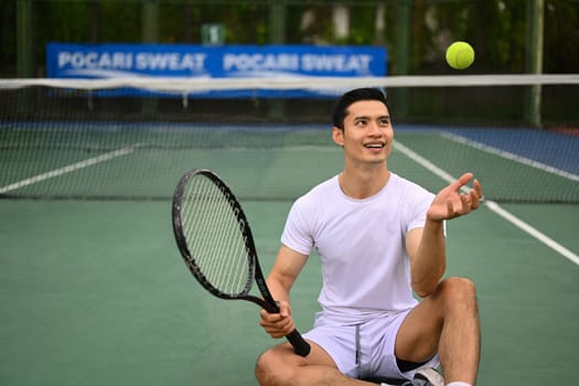 Happy sportive asian man with racket throwing tennis ball in air, resting on tennis court. Exercise, sports and wellness concept.