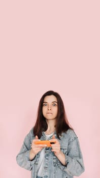 Portrait of a young woman in casual denim jacket with smartphone playing game, isolated on pink background conveying hesitancy and insecurity. Vertical photography