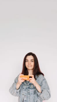 Indecisive and Insecure: Young Woman in Denim Jacket Playing Smartphone Game, Isolated on Gray Background. Emphasizing Uncertainty and Doubt. Vertical photography