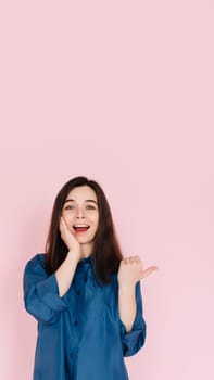 Young woman expressing amazement and joy, touching cheek and pointing finger at empty space, with a big toothy smile on pink isolated background. Vertical photography.