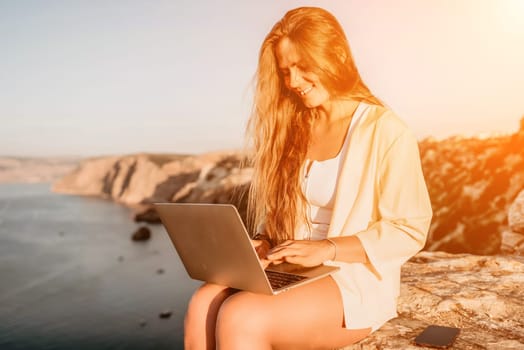 Successful business woman in yellow hat working on laptop by the sea. Pretty lady typing on computer at summer day outdoors. Freelance, travel and holidays concept.