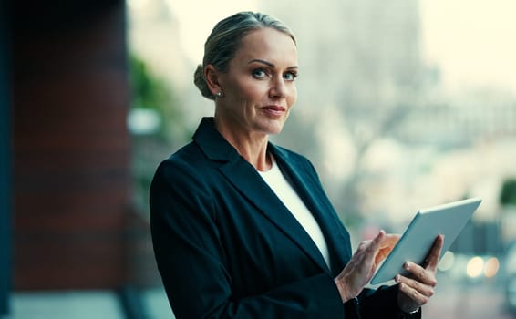 It keeps me current and connected. Portrait of a mature businesswoman standing outside on the balcony of an office and using a digital tablet