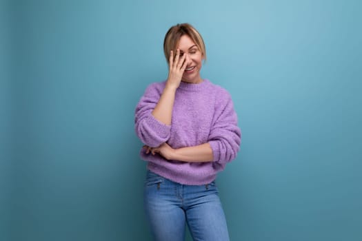 cute blond young woman in a lilac sweater stands embarrassedly on a bright background with copy space.