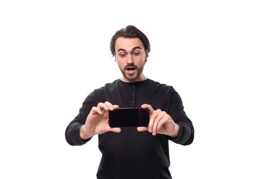 handsome european macho man with black hair and beard with an earring in his ear in surprise shows the screen of a smartphone horizontally standing on an isolated white background with copy space.