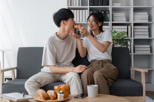 Young couples spend holidays in the living room. The young man was wearing comfortable clothes, showing his face, satisfied with the taste of snacks. Eating croissant on the sofa.
