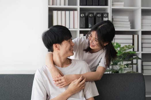 Happy young asian couple relax on cozy sofa at home hugging look at each other, enjoy weekend at home.