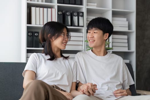 Happy young asian couple relax on cozy sofa at home hugging look at each other, enjoy weekend at home.