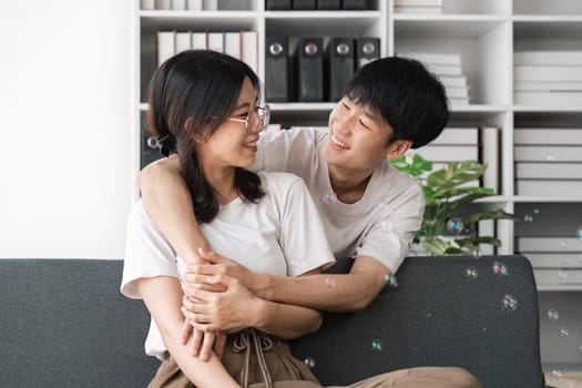 Happy young asian couple relax on cozy sofa at home hugging look at each other, enjoy weekend at home.