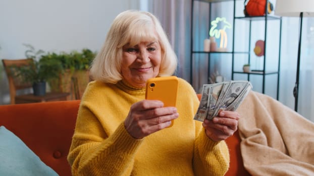Planning family budget. Senior grandmother counting money cash, use smartphone calculate domestic bills at home. Joyful elderly woman satisfied of income and saves money for planned vacation, gifts