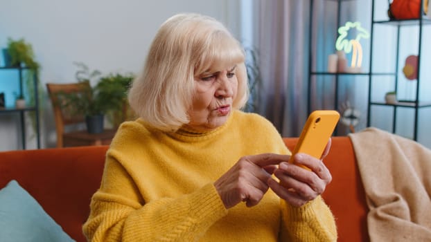 Sad displeased senior grandmother use smartphone typing browsing, loses becoming surprised sudden lottery results, bad news, fortune loss, fail, deadline, virus, debt. Elderly woman at home on couch