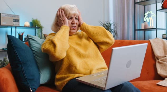 Displeased senior grandmother use laptop notebook typing browsing working, loses becoming surprised sudden lottery results bad news fortune loss fail computer virus. Elderly woman at home on couch