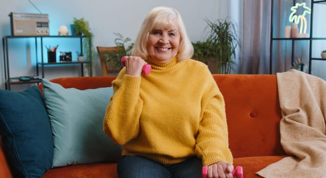 Senior grandmother woman doing dumbbells weight lifting fitness exercises at home apartment indoors. Mature old pensioner workout training strength practicing effort in room on sofa. Sport motivation