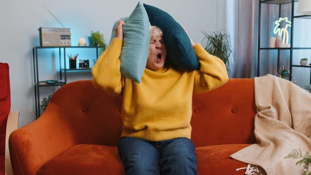 Repair work at neighbours. Irritated old woman relaxing on couch cover ears with pillows annoyed by noisy neighbors suffer from headache wish silence. Thin walls at home flat without sound insulation