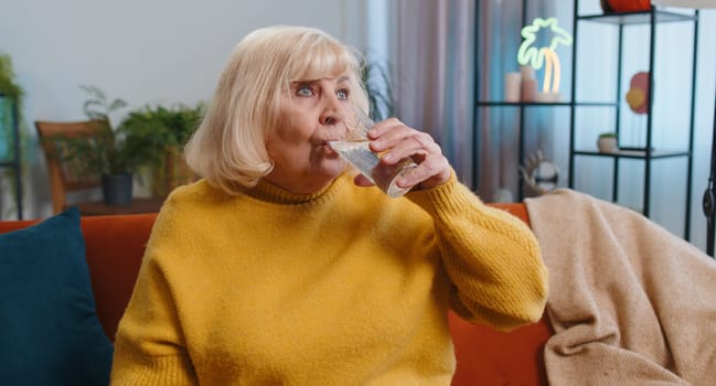 Thirsty grandmother holding glass of natural aqua make sips drinking still water preventing dehydration sits at home room. Senior old woman with good life habits, healthy slimming, weight loss concept
