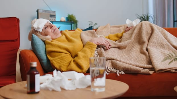 Grandmother ill suffering from cold or allergy pain lying on couch in living room at home. Sick elderly woman with towel on head blows sneezes wipes snot into napkin. Coronavirus quarantine pandemic
