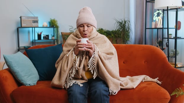 Sick senior grandmother woman in plaid shivering from cold on sofa drinking hot tea in unheated apartment without heating due debt. Unhealthy ill elderly pensioner feeling discomfort try to warming up