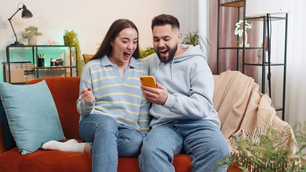Family couple man woman use mobile smartphone found out great big win good news celebrate lottery jackpot lucky victory, playing game shopping online at home. Husband and wife together on sofa in room