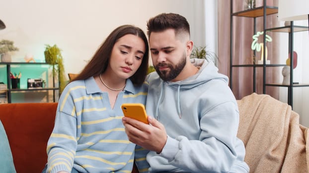 Young family married couple man woman use smartphone typing browsing, loses becoming surprised sudden lottery results, bad news, fortune loss, fail at home. Husband and wife together on sofa in room