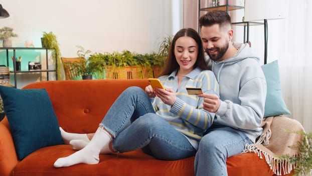 Family married couple man woman using credit bank card and smartphone while transferring money, purchases online shopping, order food delivery at home. Husband and wife together on sofa in living room
