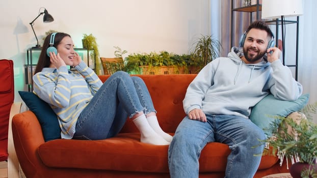 Happy overjoyed young family married couple man woman in wireless headphones dancing on couch at home listening favorite energetic disco rock n roll music. Husband and wife together weekend activities