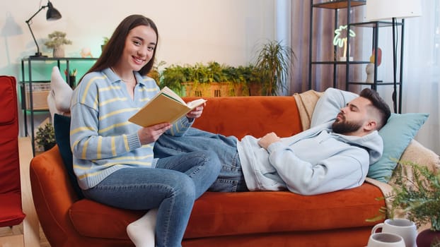 Woman reading story from book while displeased tired boyfriend man falling asleep and snoring. Young family couple at home. Husband wife together on sofa in living room. Hobby leisure activities