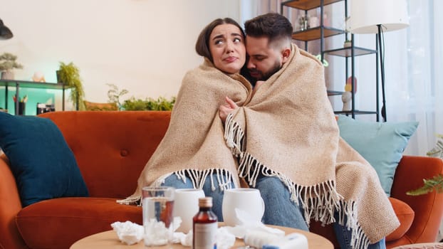 Young ill family married couple man woman suffering from cold or allergy on home sofa in living room. Sick husband wife together blows sneezes wipes snot into napkin. Coronavirus quarantine, pandemic