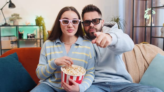 Excited young family marriage couple man woman eating popcorn and watching interesting tv serial, sport game, film, online social media movie content at home. Husband and wife together on sofa in room