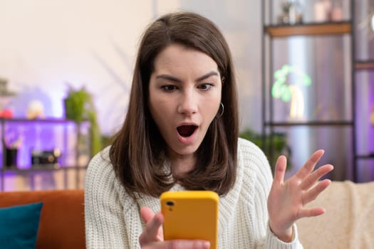Oh my God Wow. Excited happy joyful winner young woman use smartphone typing browsing found out great win good news celebrate victory at home room apartment. Pretty girl with mobile phone on couch