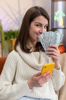 Planning family budget. Smiling young woman counting money cash, use smartphone calculate domestic bills at home room. Joyful girl satisfied of income and saves money for planned vacation, gifts