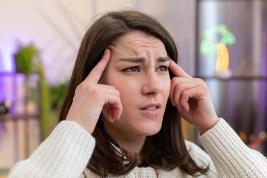 Displeased young woman rubbing temples to cure headache problem, suffering from tension and migraine, stress grimacing in pain high blood pressure at home room apartment. Girl on couch. Lifestyles