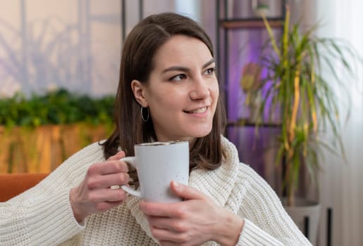 Portrait of attractive smiling young woman drinking a cup of warm coffee or herbal tea at home living room apartment in the morning. Pretty girl lying on couch enjoying comfortable relaxing alone