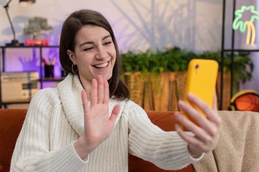 Young woman making online selfie video call talking waving hand hello, looking at camera at home apartment indoors. Communication, meeting. Brunette pretty girl blogger making smartphone conversation