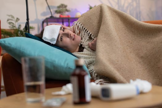 Young ill woman suffering from cold or allergy sitting on sofa in living room at home. Sick girl wear hat wrapped in plaid blows sneezes wipes snot into napkin. Coronavirus quarantine, pandemic