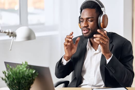 An African-American man sits at his desk in front of his laptop, wearing headphones and chatting on a video call, listening to music. The concept of student business training and online work. High quality photo