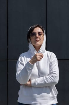 Portrait Mature Brunette Woman With Hood On Head, Wears White Hoodie and Sunglasses Looks Straight, Dark Background Wall. Sportive Confident 40 Yo Beautiful Caucasian Woman Outdoor. Vertical Plane.